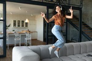 Happy young Caucasian woman in wireless headphones enjoying music, dancing and jumping on sofa at